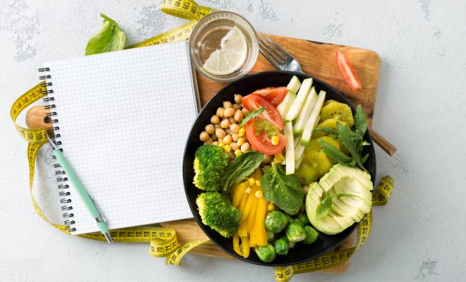 Vegan healthy balanced diet concept. Vegetarian buddha bowl with blank notebook and measuring tape. hickpeas, broccoli, pepper, tomato, spinach, arugula and avocado in plate on white background. Top view